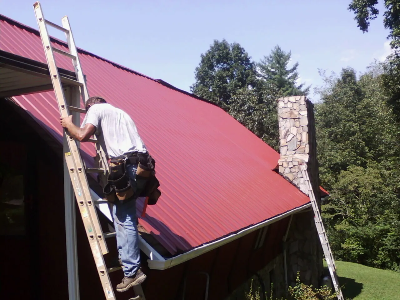 Roofer on a ladder beside a red metal roof and stone chimney — Soffit & Fascia Repair by Roofing Brothers in Bentonville